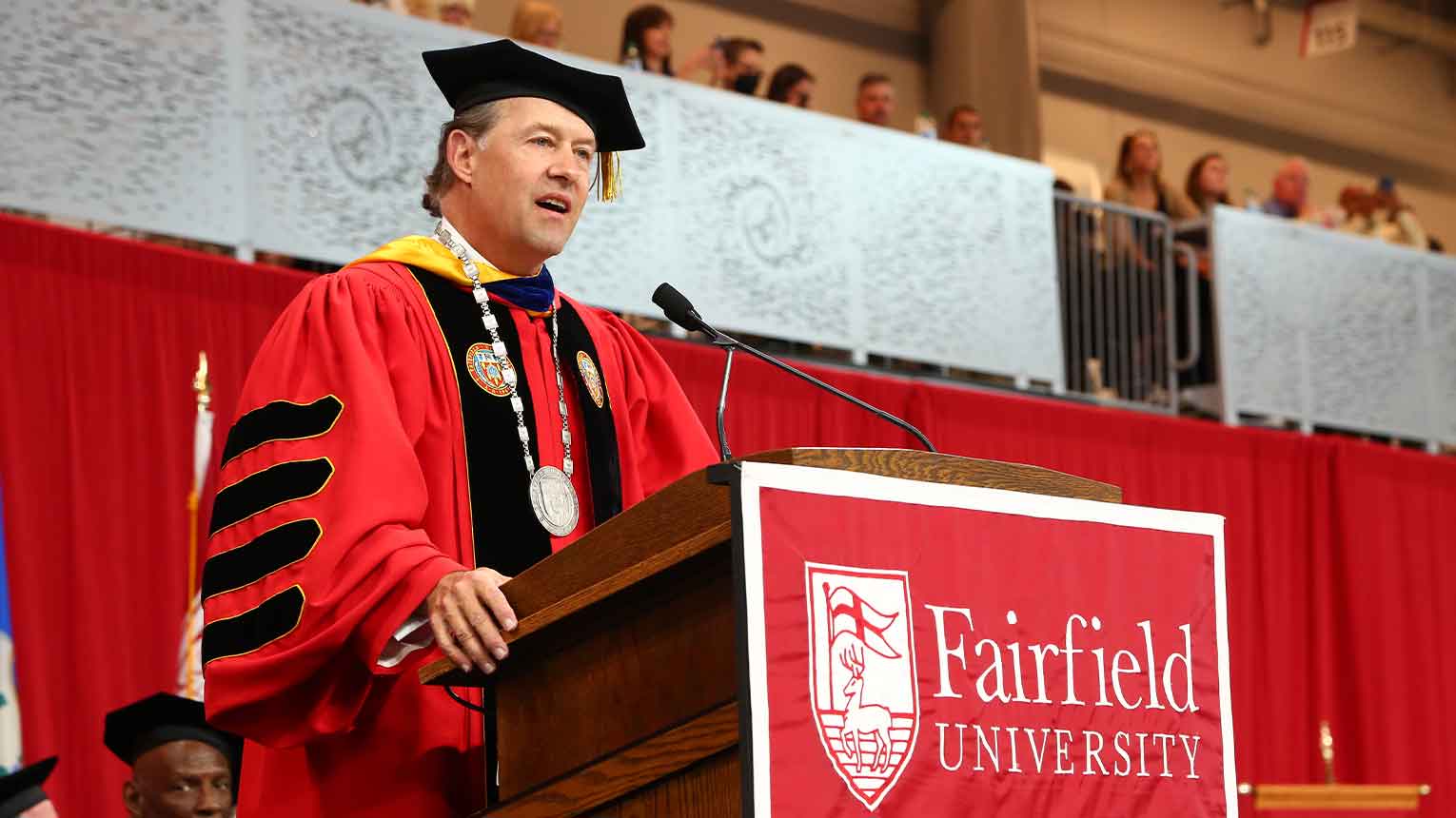 Mark Nemec in red commencement attire addressing audience at Graduate Commencement.