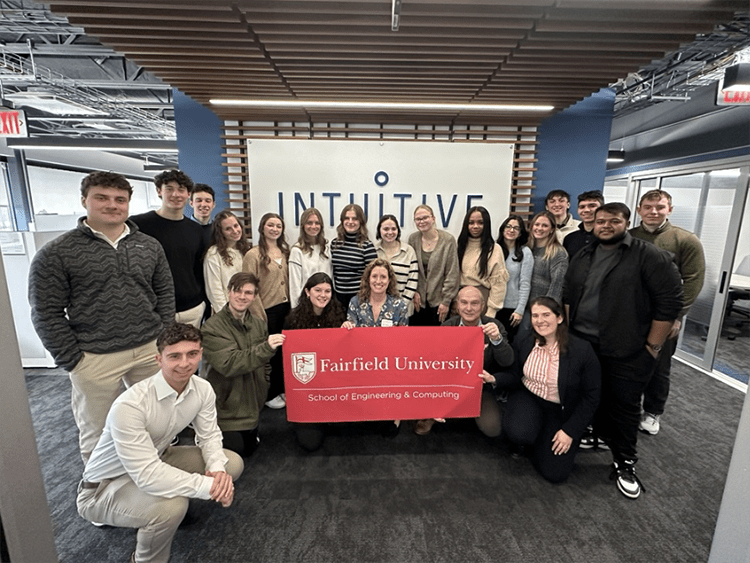 Fairfield students posing for a group photo with a Fairfield U-branded banner in front of the indoor Intuitive Surgical logo wall.