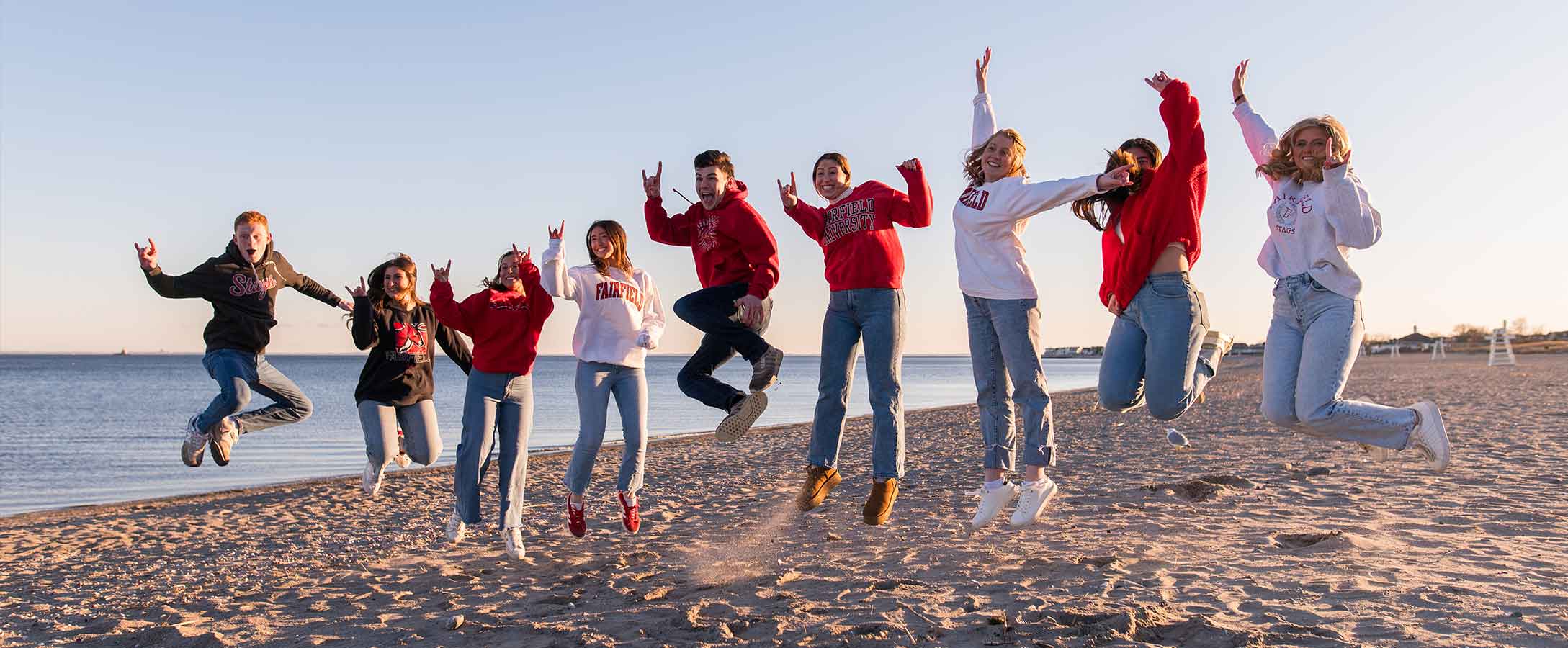 A group of fairfield students jumps in the air on the beach while the sun is setting.