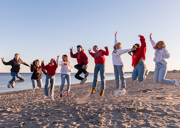 A group of fairfield students jumps in the air on the beach while the sun is setting.