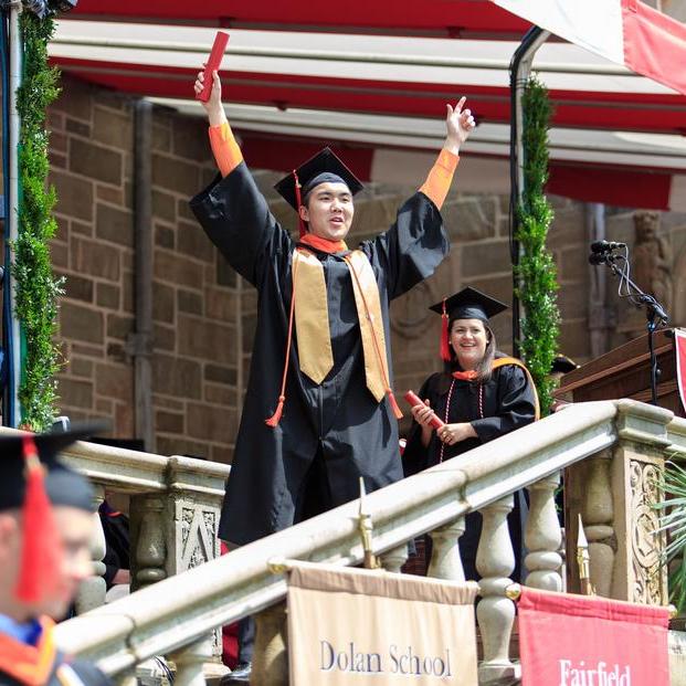 A student in a cap and gown holding a diploma tube holds both his hands up.