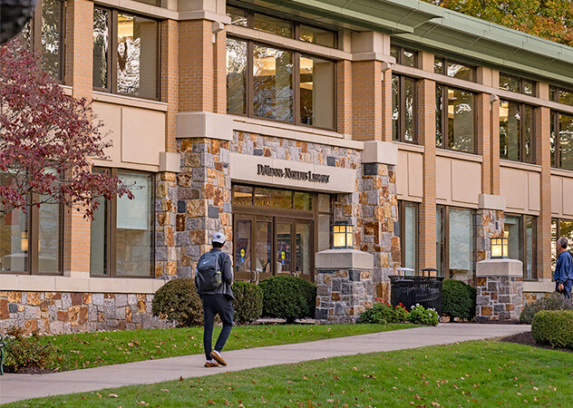 A student walking on the sidewalk, with a building visible in the background, showcasing campus life.