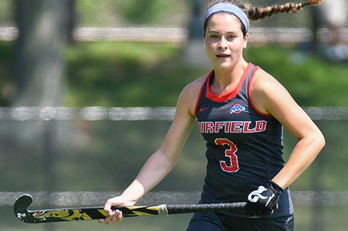 A field hockey player in a black jersey with red letters that read “Fairfield 3” is in motion.