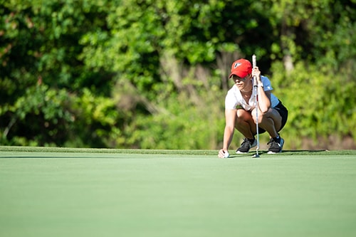 A golf player in a white shirt and red Fairfield hat holds a golf club and moves a golf ball.