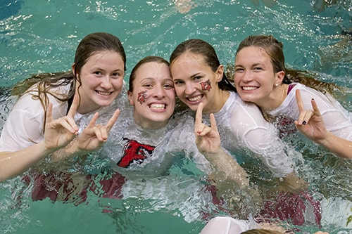 A group of four students in white shirts with a red “F” pose for a photo in a pool. 