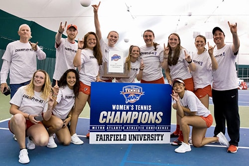 A group of people pose in front of a sign that reads “Women’s Tennis Champions, Metro atlantic Athletic Conference, Fairfield University”. 