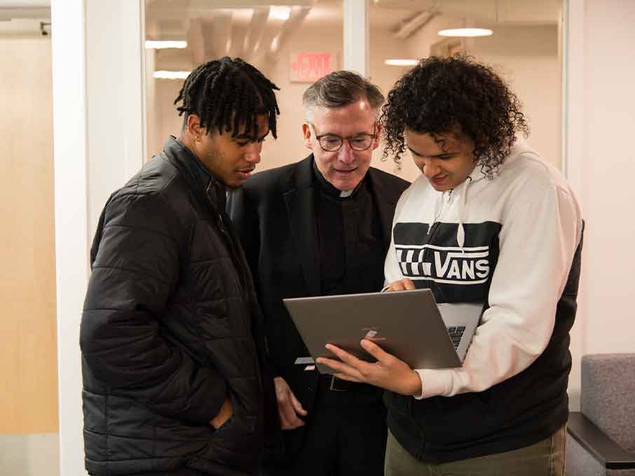 a priest looks at a laptop with two students.