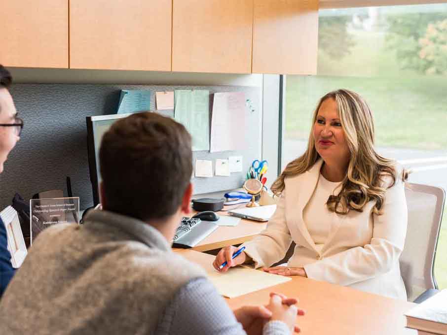 A professor sits at her desk and addresses two students.