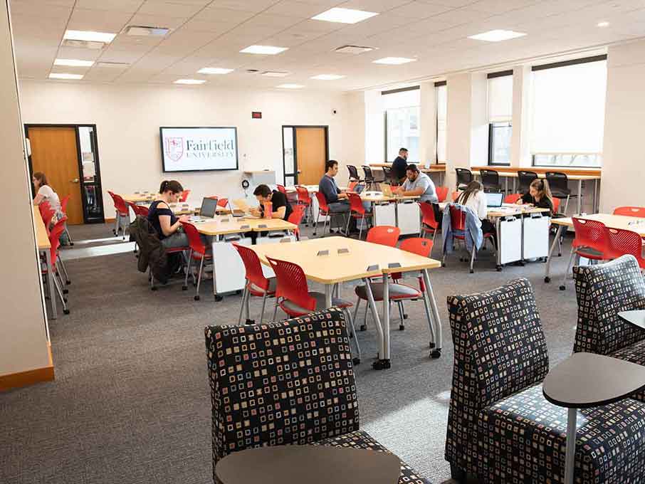 A room full of tables and chairs with some students working in front of a screen that reads “Fairfield University”