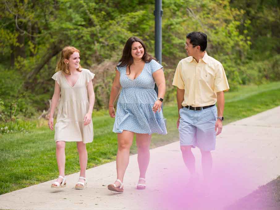 People walk outdoors on a concrete path, slightly obscured by a blurry purple flower on the bottom.