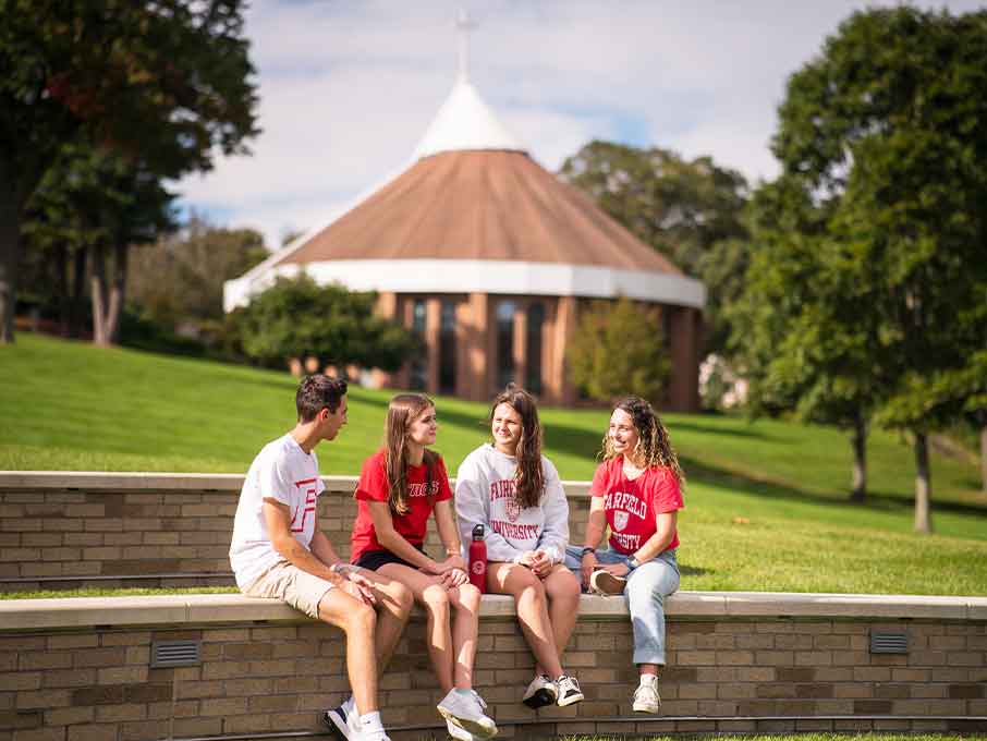 Four students seated on a brick wall, with a church in the background, enjoying a moment together outdoors.