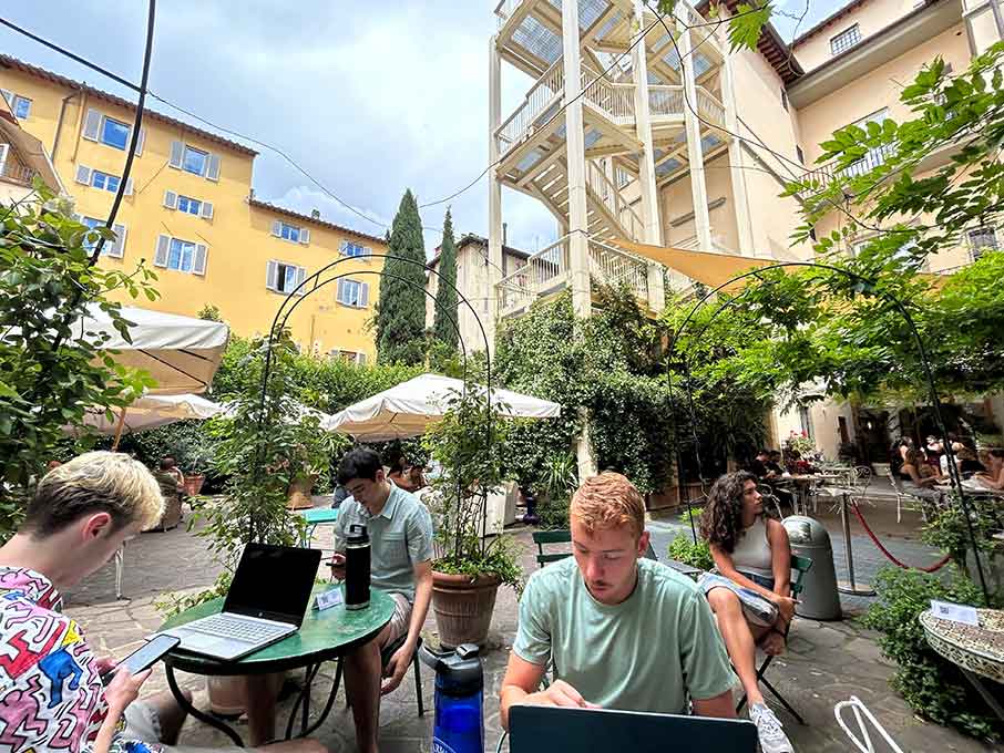 A group of students sit at tables in front of their open laptop in a courtyard.