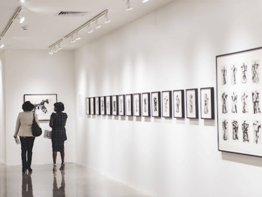 Two people stand at the end of a gallery with paintings hanging on a white wall.