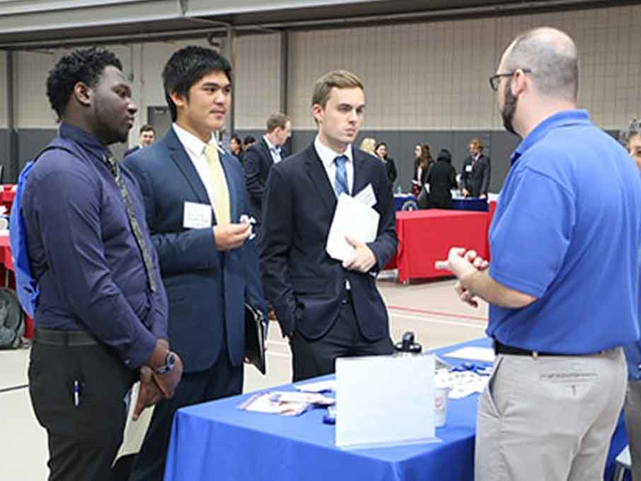 Students wearing suits stand around a table at a career fair listening to a man in a blue collared shirt speak to them. 