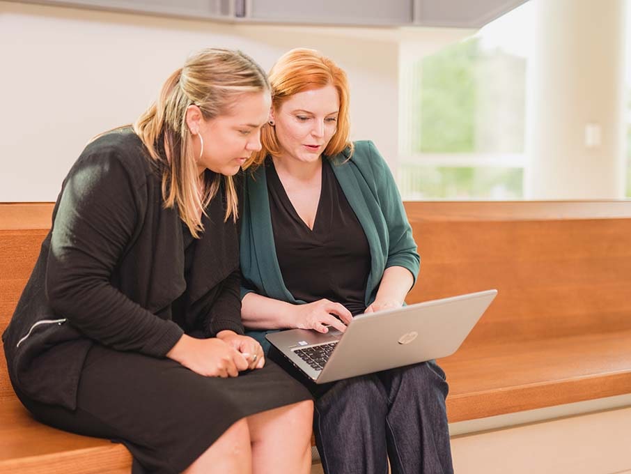 A student sits with Professor Gayle Alberda on a wooden bench and look at a laptop.