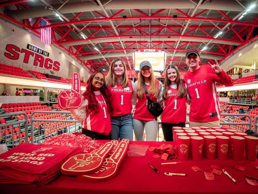 A group of students wear Fairfield jerseys with foam fingers in front of other Fairfield merch. 