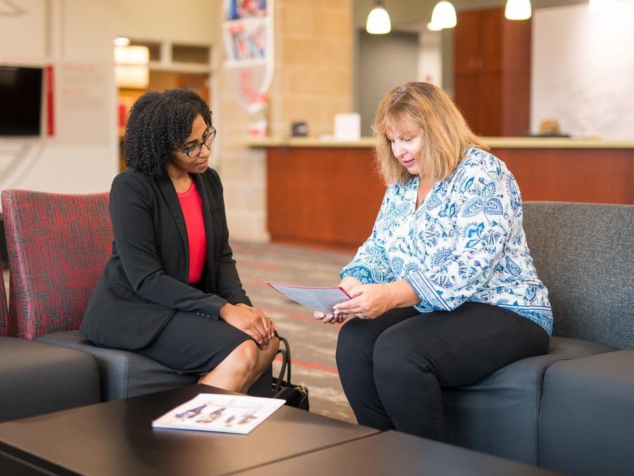 A graduate student in a suit speaks with a staff member who shows her a document. They are seated in a lounge area in discussion.