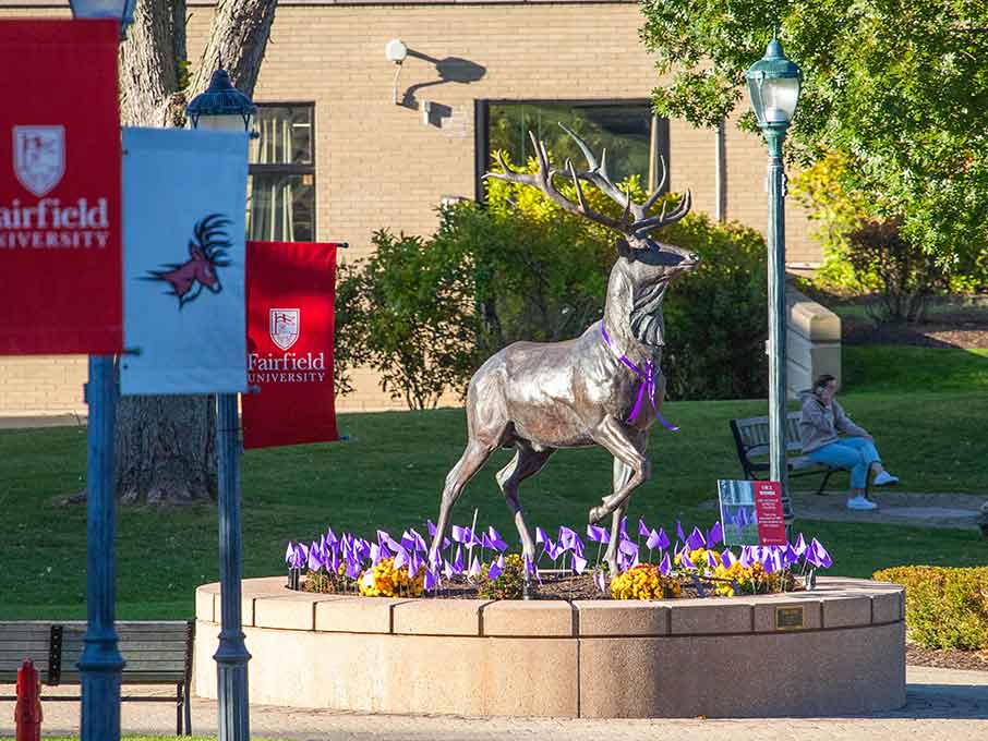 The Stag Statue at the end of a path with Fairfield University lightpoles and flags on the left side.