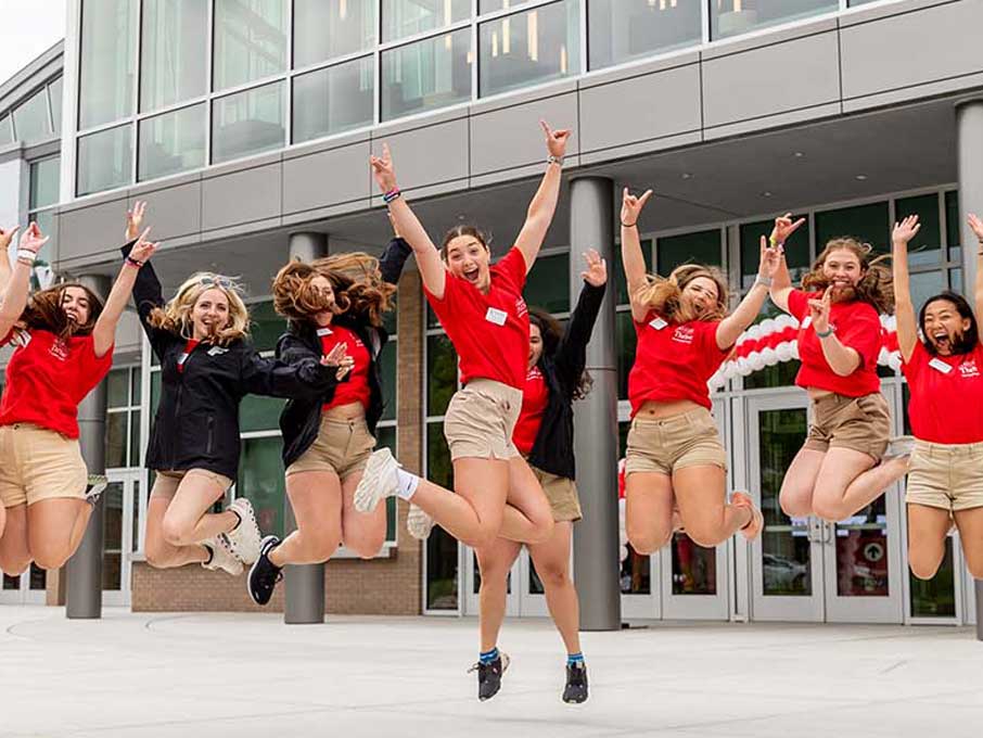 Students jumping in the air and throwing their hands up in front of a university building. 