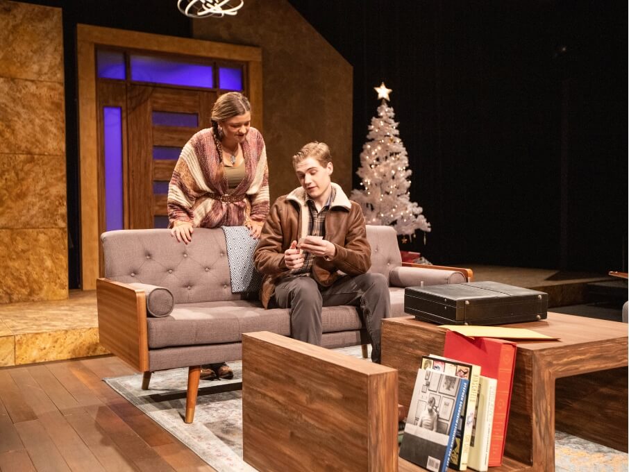 One actor sits on a couch on set while an actress stands behind him. 