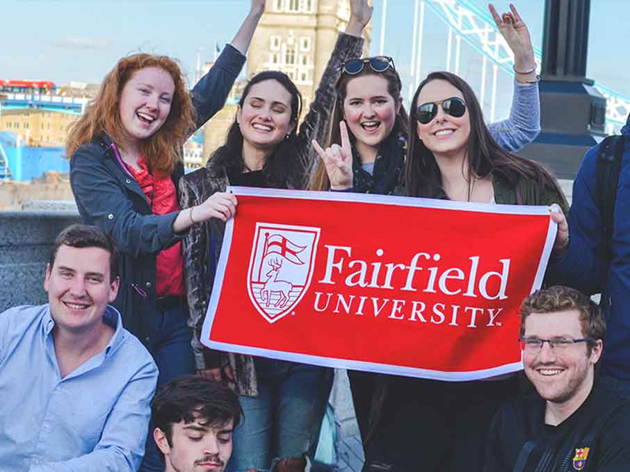 A group of Fairfield students holds a red banner that reads Fairfield University in front of the Tower Bridge in London.