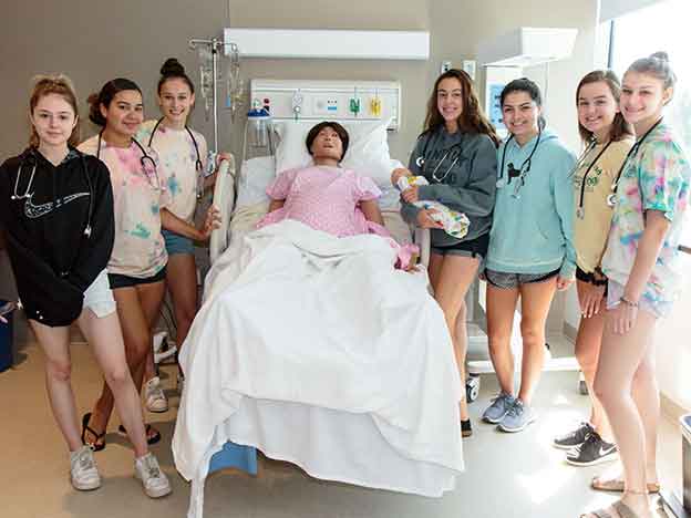 A group of students smile gathered around a manikin in a hospital bed.