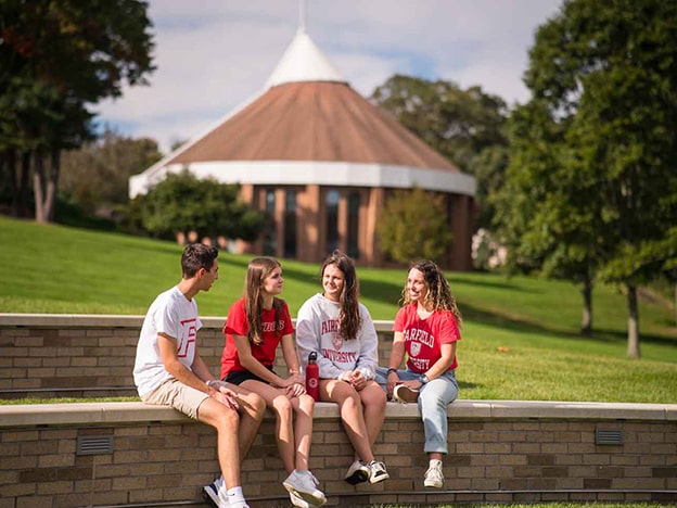 Three students sit on a wall with the library lawn and a chapel in the background