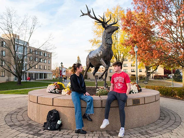 Two students sit on wall next to Stag Statue with backpacks on the ground.