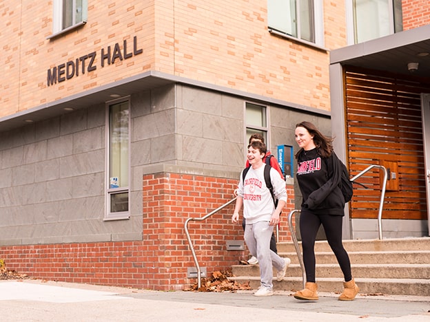 Two students walk down the steps of a building that is marked “Meditz Hall”.