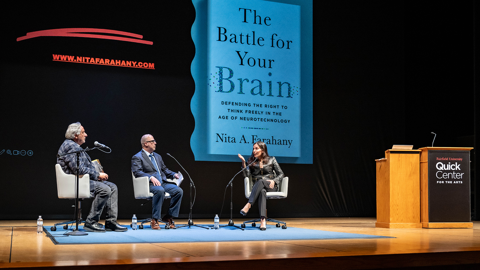 Three individuals seated on chairs before a stage displaying a banner that reads "The Battle for Your Brain."