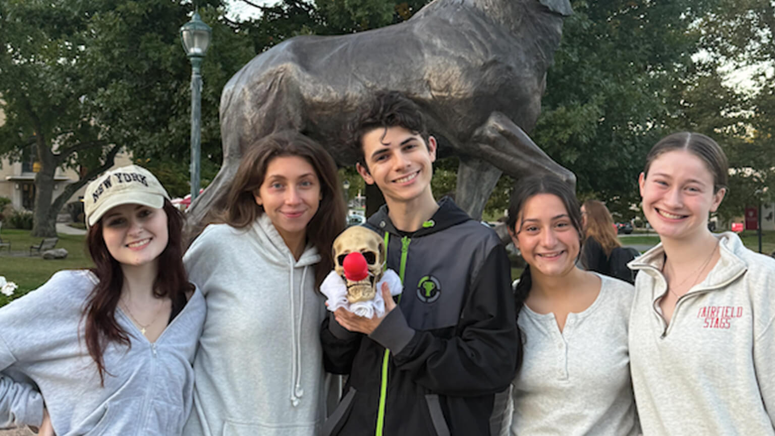 Five young people smile for a photo beside the Stag statue on Fairfield University's campus in a sunny outdoor setting. 