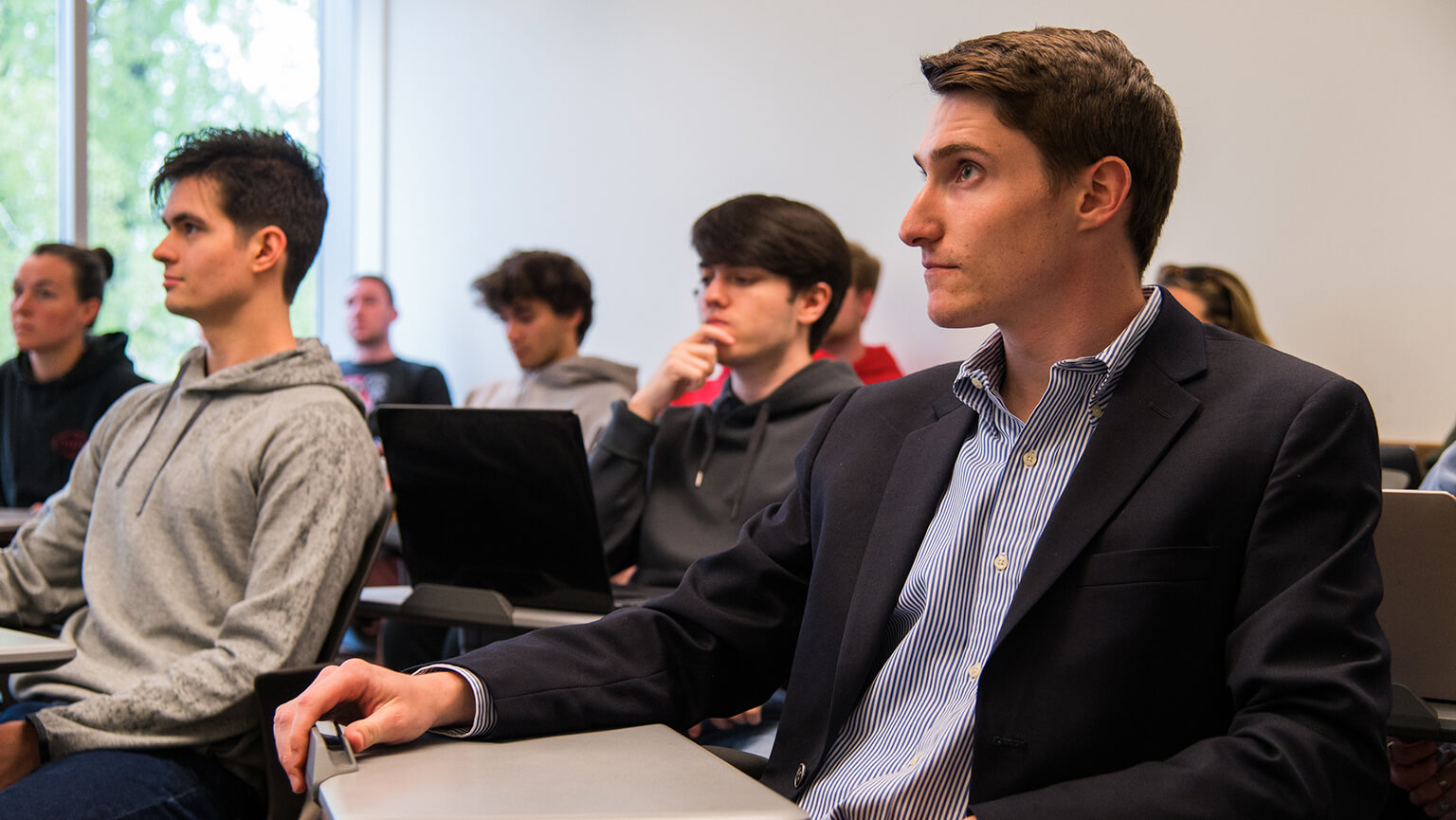 A man seated in a classroom surrounded by fellow students, engaged in a learning environment.