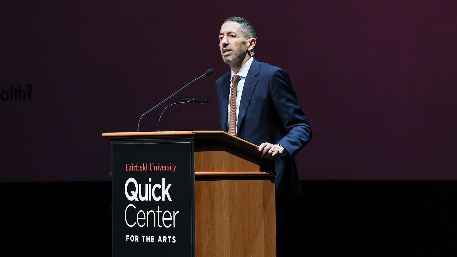 A man wearing a suit and tie stands at a podium, poised to speak, with a serious demeanor and engaged audience in view.