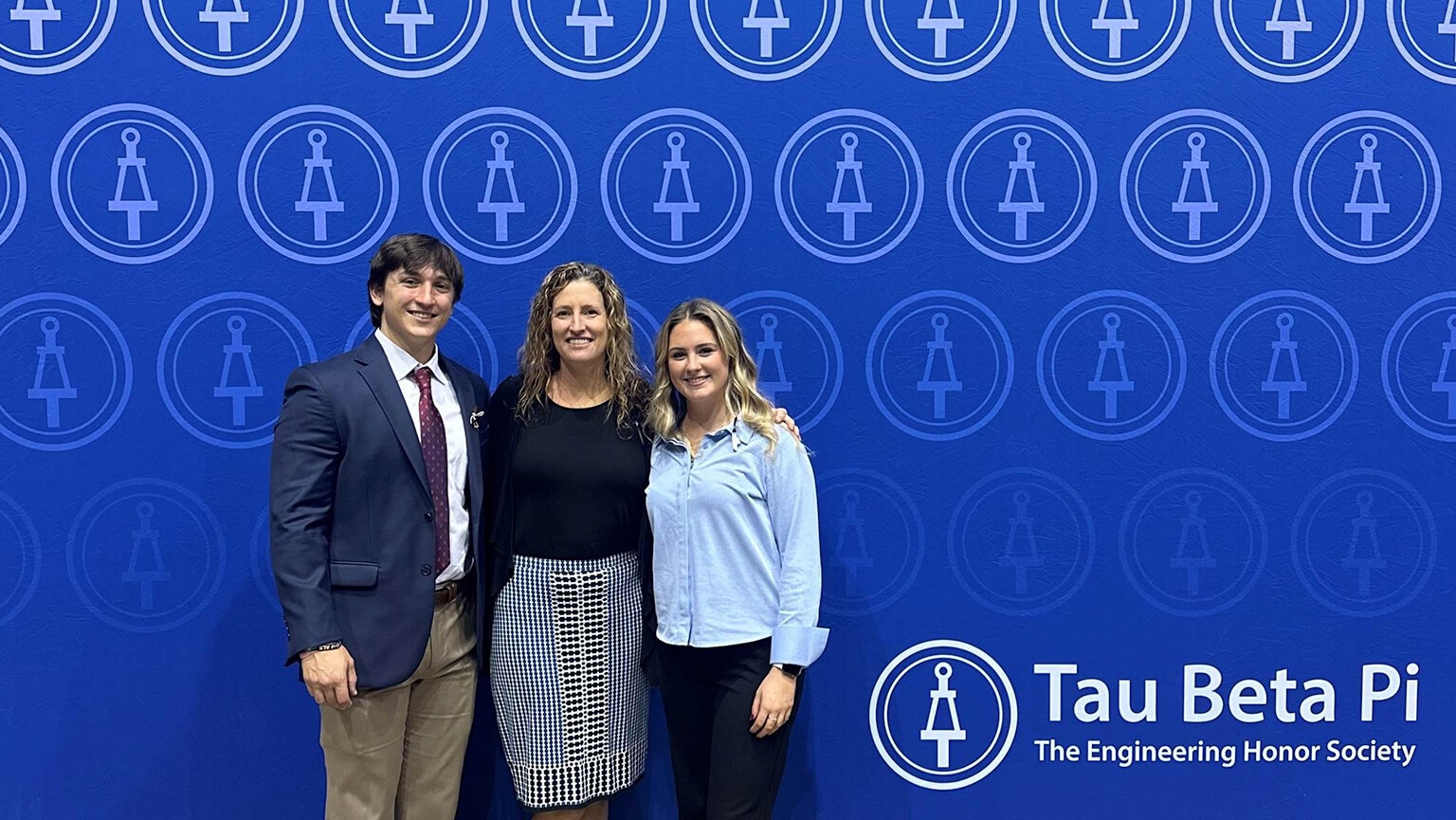 Three individuals pose in front of a wall displaying the words "Tau Beta Pi," showcasing their affiliation with the organization.