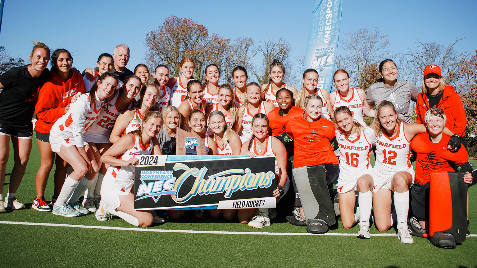 The women's field hockey team stands together, smiling for a group photo on the field, showcasing their championship banner.