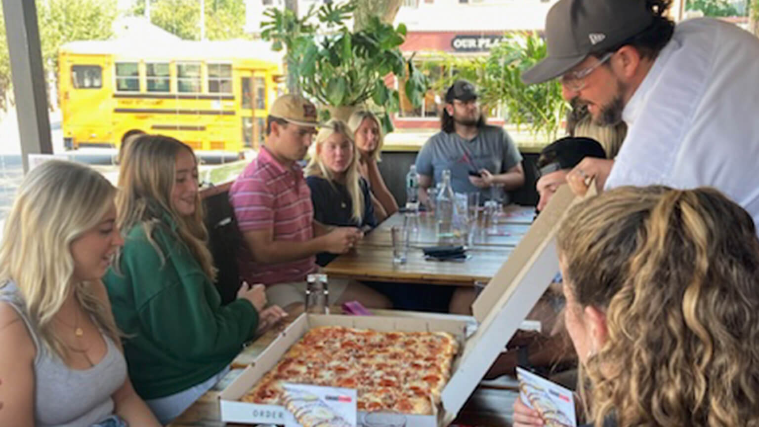 A group of young adults dining at a restaurant as a server presents a large pizza to them.