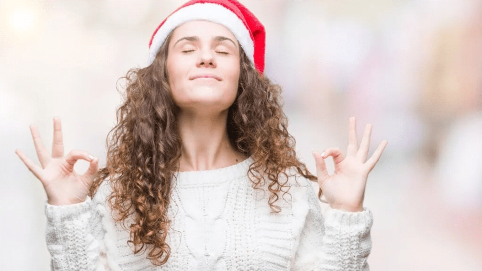 Curly-haired woman in a Christmas Santa hat appearing to meditate while smiling.