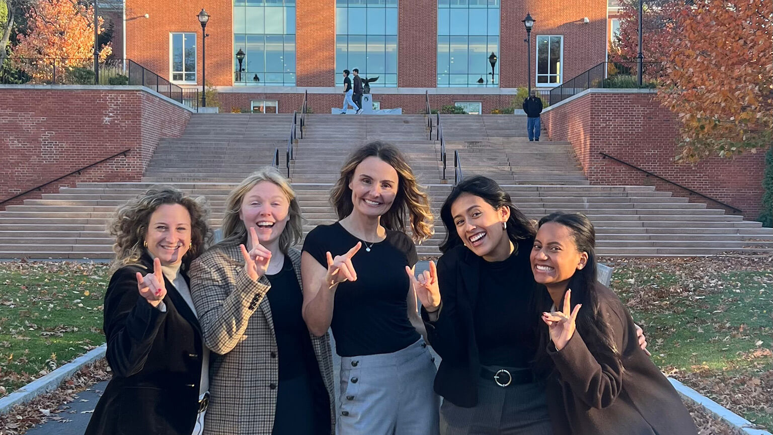 Female Marriage & Family Therapy students posing with the 'Stags Up' hand sign outside the main Bentley University building.