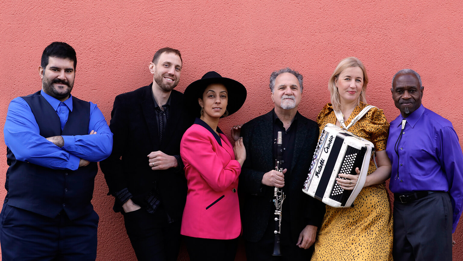 A diverse group of individuals stands together, smiling, with one person playing an accordion among them.