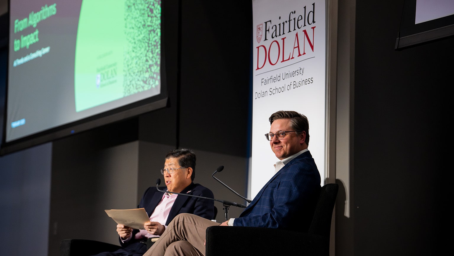 Two men sitting and presenting on stage in an engaging discussion as part of a forum.