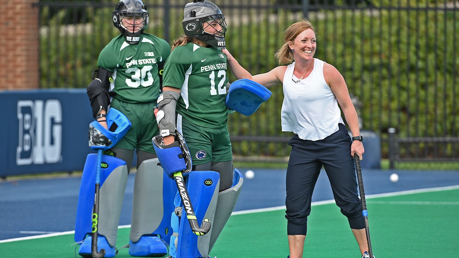 Field Hockey coach smiling as she poses for a candid photo with two of her geared-up student-athletes.