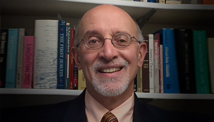 John E. Thiel, PhD sitting in front of bookshelf. 