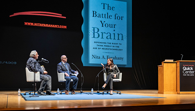 Three individuals seated on chairs before a stage displaying a banner that reads "The Battle for Your Brain."