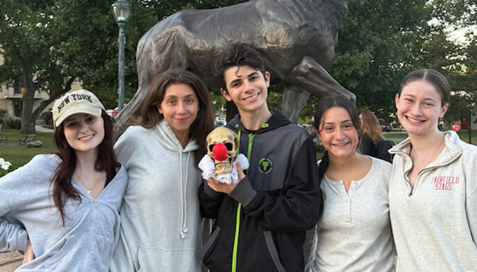 Five young people smile for a photo beside the Stag statue on Fairfield University's campus in a sunny outdoor setting. 