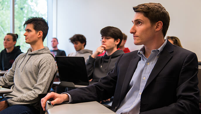 A man seated in a classroom surrounded by fellow students, engaged in a learning environment.