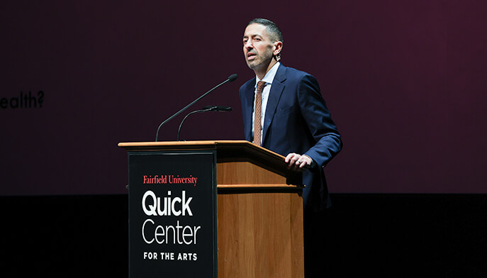 A man wearing a suit and tie stands at a podium, poised to speak, with a serious demeanor and engaged audience in view.