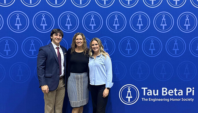 Three individuals pose in front of a wall displaying the words "Tau Beta Pi," showcasing their affiliation with the organization.