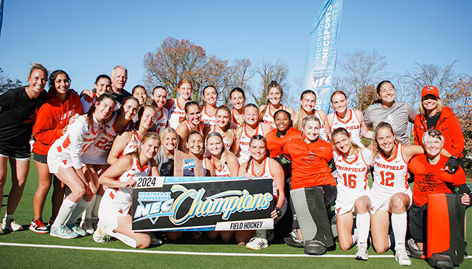 The women's field hockey team stands together, smiling for a group photo on the field, showcasing their championship banner.