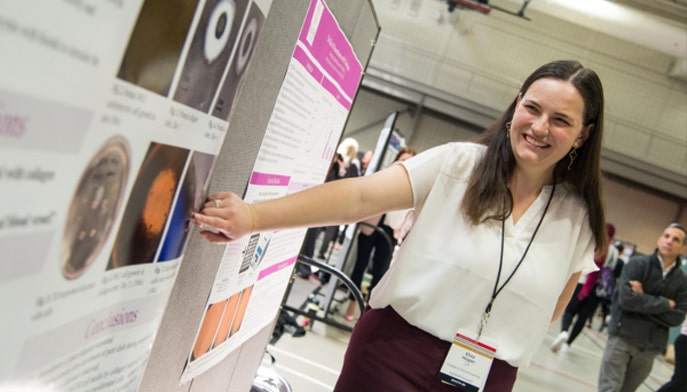 Student pointing at her research work and explaining it to group of others.