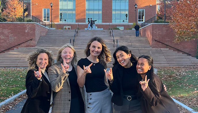 Female Marriage & Family Therapy students posing with the 'Stags Up' hand sign outside the main Bentley University building.